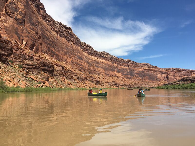 The image shows a scenic view of a river with two canoes paddling through the calm water. Towering red rock cliffs rise on the left side of the river, contrasting with the blue sky and scattered white clouds above. Green vegetation lines the riverbanks, adding a touch of life to the arid landscape. The overall scene evokes a sense of adventure and tranquility in a natural setting.